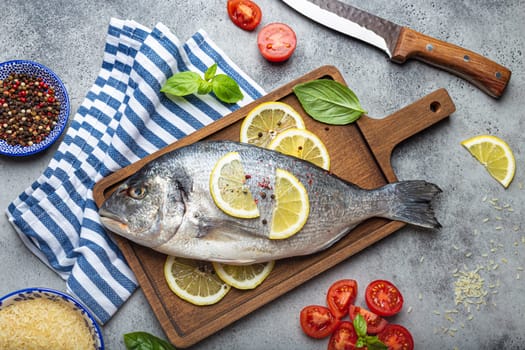 Raw fish dorado with ingredients lemon, fresh basil, cut cherry tomatoes, uncooked rice on wooden cutting board with knife on rustic stone background top view, cooking healthy fish dorado concept