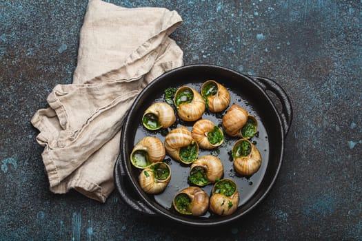 Escargots de Bourgogne Cooked Snails with Garlic Butter and Parsley in black cast iron pan on rustic stone background top view, traditional French Delicacy .