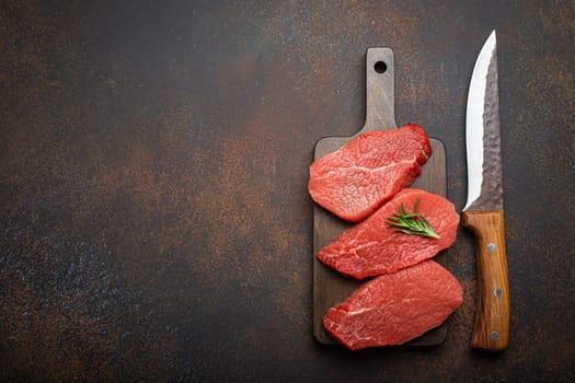 Three raw uncooked meat beef top sirloin steaks on wooden cutting board with rosemary on wooden cutting board with knife on dark rustic background from above, preparing meal with meat, copy space