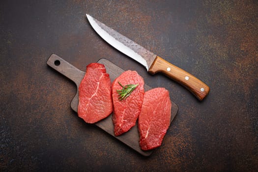 Three raw uncooked meat beef top sirloin steaks on wooden cutting board with rosemary on wooden cutting board with knife on dark rustic background from above, preparing meal with meat