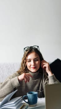 Stylish woman enjoying online education with a cup of coffee at the kitchen table, wearing headphones and writing notes while watching a webinar on her laptop.