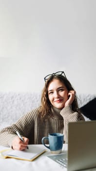 Happy student studying at home, doing homework and preparing for exams in front of laptop, sitting at kitchen table with pen and notebook, smiling at camera.