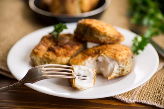 A piece of hake fish fried in a crispy crust, in a plate on a wooden table.