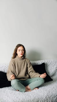 Young woman sitting in lotus position on the sofa with her hands on her knees, meditating, trying to relax her mind and relieve stress.