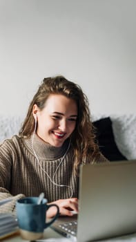 Student video chatting with friends, taking a break from studying at home. Young woman in headphones sitting at kitchen table with laptop and coffee, smiling while chatting with friends on video call.