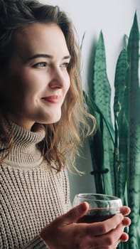 A serene young woman savors a hot cup of tea while admiring the stunning cityscape from a window, taking a peaceful moment to herself to unwind and appreciate the beauty around her