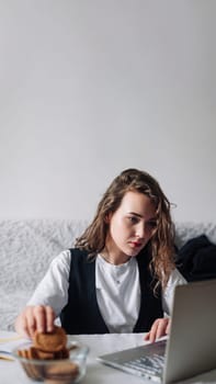 Young woman watching series or podcasts, looking at the screen with interest, holding cookies in hand, sitting at the table with a glass jar and laptop, smiling happily. Unconscious Eating Disorder.