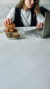 Unconscious eating. Bad habits. Selective focus of hand of concentrated female working on laptop at the kitchen table, taking unintentionally and automatically cookies from a transparent glass jar.