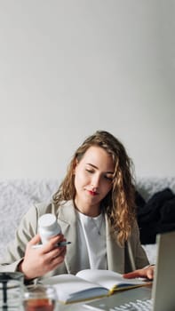 Nutritionist writing article about effects of supplement deficiency on human health, working on laptop at kitchen table with bottle of vitamins nearby.