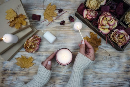 A woman lights an aromatic candle. There are dried roses in a box on the table and yellow maple leaves are lying. . High quality photo