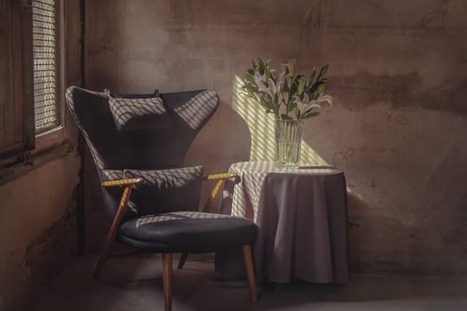 A Wooden Upholstered Chair and the Bouquet of Flowers in a Glass Vase on Table in the Room with Bare Cement Wall. They are affected by the Sunbeam Shining through Steel Mesh Window in the Afternoon. Space for text, Selective focus.
