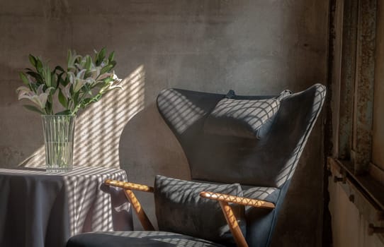 A Wooden Upholstered Chair and the Bouquet of Flowers in a Glass Vase on Table in the Room with Bare Cement Wall. They are affected by the Sunbeam Shining through Window in the Afternoon. Space for text, Selective focus.