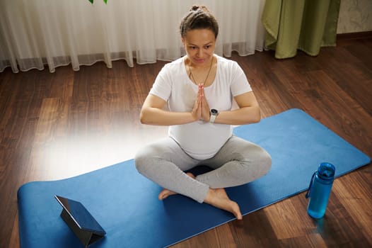 Happy smiling pregnant woman sitting on mat with online yoga class on digital tablet while training at home. Expecting future mother enjoying healthy lifestyle. Fitness during pregnancy. Overhead view