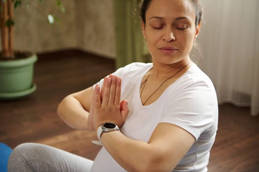 Closeup shot. Gorgeous adult pregnant woman, gravid female meditating at home. Expectant lady sitting in lotus position with clasped hands, practicing yoga for healthy pregnancy. Maternity. Childbirth