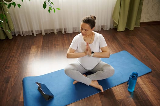View from above of pregnant woman sitting in comfortable lotus position on a fitness mat, keeping her hands palms together, practicing online pregnancy yoga, watching video classes for gravid mothers