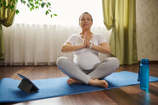 Pregnant gravid woman sitting barefoot on a fitness mat, keeping hands palms together, deeply breathing while practicing online pregnancy yoga, watching video class on digital table at home. Maternity