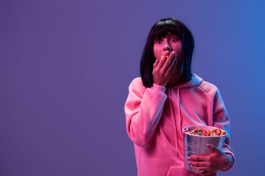 Shocked scared awesome brunet woman in pink hoodie eat popcorn look aside posing isolated in blue violet color light studio background. Neon party Cyberpunk Cinema concept. Copy space