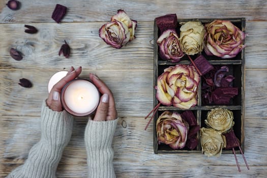 Women's hands hold a burning candle. Books in craft covers are laid out on the table, there is a wooden box with dried rose flowers and yellow autumn leaves.