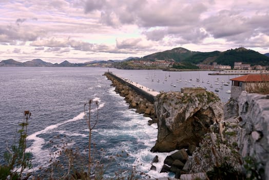 Castro Urdiales breakwater and fishing harbour in Spain. Moving sun and clouds, small boats, rocks, calm sea, mountains and vegetation.