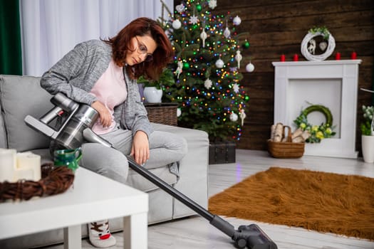 In the living room, a girl sits on the sofa and relaxes while vacuuming the floor. The living room decorated for Christmas.