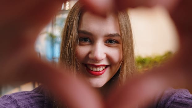 I love you. Happy young woman at home living room couch makes symbol of love, showing heart sign to camera, express romantic feelings express sincere positive feelings. Charity, gratitude, donation