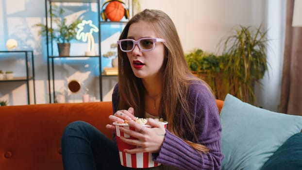 Excited woman in 3D glasses sits on couch eating popcorn snacks and watching interesting TV serial sport game, film online social media movie content at home apartment. Girl enjoying entertainment