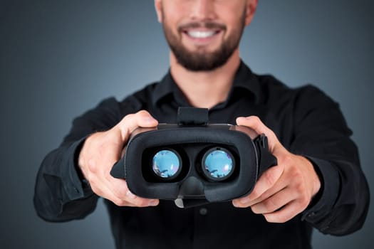 Excited young man using a VR headset and experiencing virtual reality on grey blue background. He holds glasses for viewer