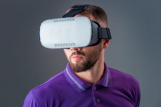 Emotional young man using a VR headset and experiencing virtual reality on grey background. A man dressed in a purple T-shirt