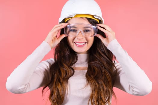 A surprised brunette in a helmet wears glasses on the pink background. Young happy architect.
