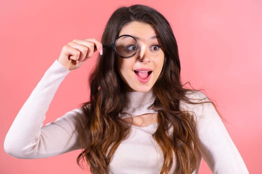 A HAppy emotional woman looks through a magnifying glass on the pink background.