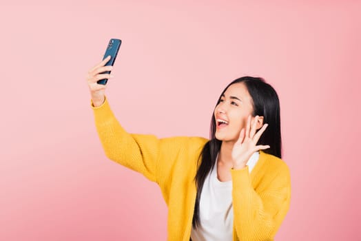 Happy Asian portrait beautiful cute young woman smiling excited making selfie photo, video call on smartphone studio shot isolated on pink background, female hold mobile phone raise hand say hello