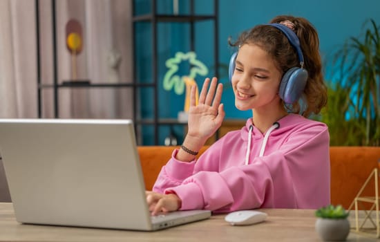 Preteen school girl looking at laptop camera, making video webcam conference call with friends or teacher, enjoying pleasant conversation, waving hello hi. Young child, kid at home workplace table