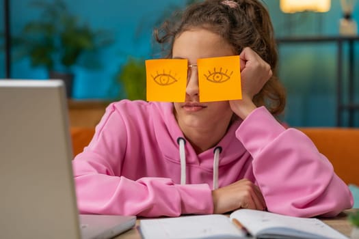Tired Caucasian preteen school girl sleeping on laptop computer with eyes stickers on face. Young lazy child, kid cheating to sleep at home workplace sitting at the table. Distance learning education