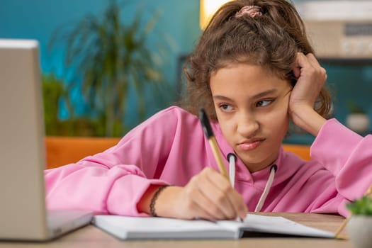 Bored tired Caucasian preteen school girl doing homework exercise online, typing on laptop, makes notes in notebook. Distance learning education. Young child kid at home workplace sitting at the table