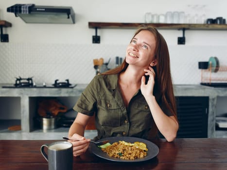 Woman with a smile eating Asian rice food in the kitchen at the wooden table, stylish interior in the background, lifestyle with phone in hand, blogger. High quality photo