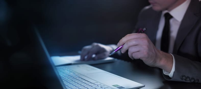 A man in a suit sitting at a laptop checks the data in a notebook, holding a pen in his hand.