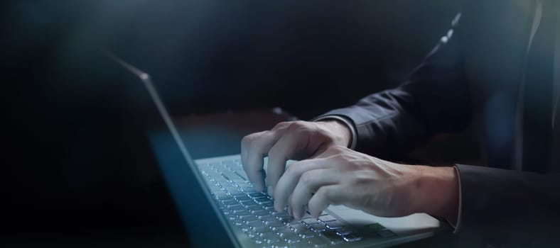On a dark background, an unrecognizable man is typing on a laptop backlit keyboard.
