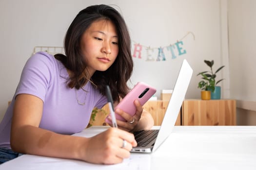 Teenage female college student doing university paper, researching with laptop and mobile phone handwriting information on piece of paper. Education concept.