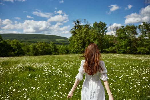 a red-haired woman in a light dress stands in a chamomile field with her back to the camera. High quality photo