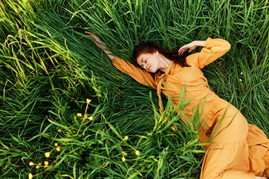 a relaxed woman enjoys summer lying in the tall green grass in a long orange dress stretched out stretching her arm forward. High quality photo