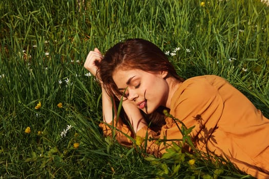 a calm woman with long red hair lies in a green field with yellow flowers, in an orange dress smiling pleasantly, closing her eyes from the bright summer sun. High quality photo