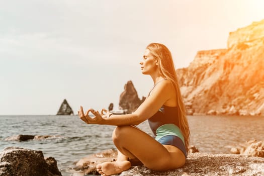 Yoga on the beach. A happy woman meditating in a yoga pose on the beach, surrounded by the ocean and rock mountains, promoting a healthy lifestyle outdoors in nature, and inspiring fitness concept