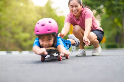 Active little girl and mom enjoy skateboarding. Cute little girl wearing helmet practicing skateboarding in park. Mother trains her daughter to skateboard. Outdoor sports for children.