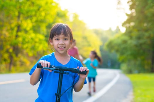 Active little girl riding a scooter on a park road on a summer day against her mother and sister in a blurred background. Family spending time together.