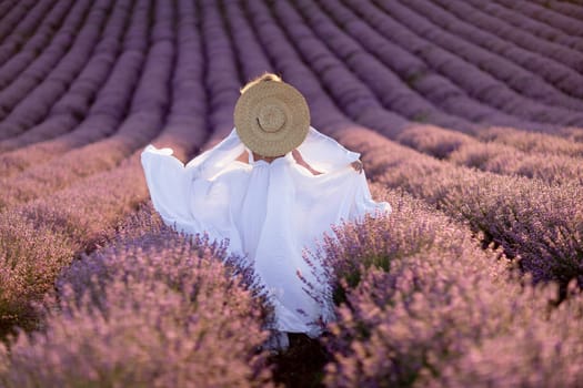 Happy woman in a white dress and straw hat strolling through a lavender field at sunrise, taking in the tranquil atmosphere