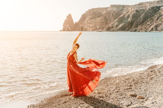 Woman red dress sea. Female dancer in a long red dress posing on a beach with rocks on sunny day. Girl on the nature on blue sky background