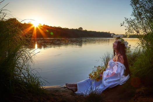 Adult mature brunette woman in a white dress, sundress and a wreath of flowers in summer by the water of river or lake in evening at sunset. Celebration of the Slavic pagan holiday of Ivan Kupala