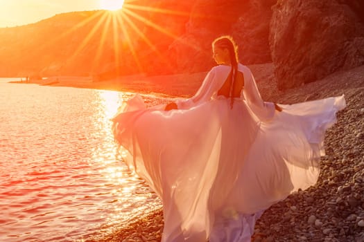 Mysterious woman silhouette long hair walks on the beach ocean water, sea nymph wind listens to the wave. Throws up a long white dress, a divine sunset. Artistic photo from the back without a face.