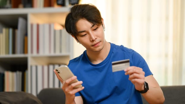 Young asian man holding credit card, paying bill with online payment via internet on smart phone.