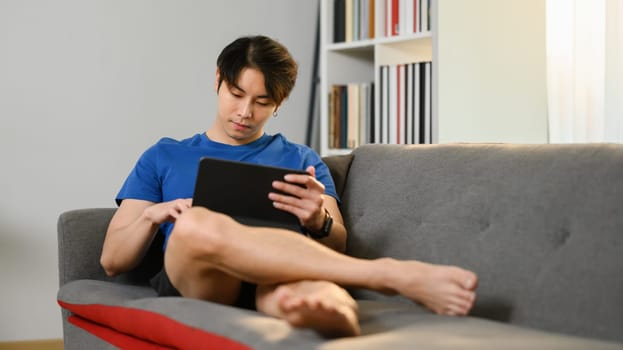 Barefoot young asian man in casual wears sitting on couch and browsing internet on digital tablet.